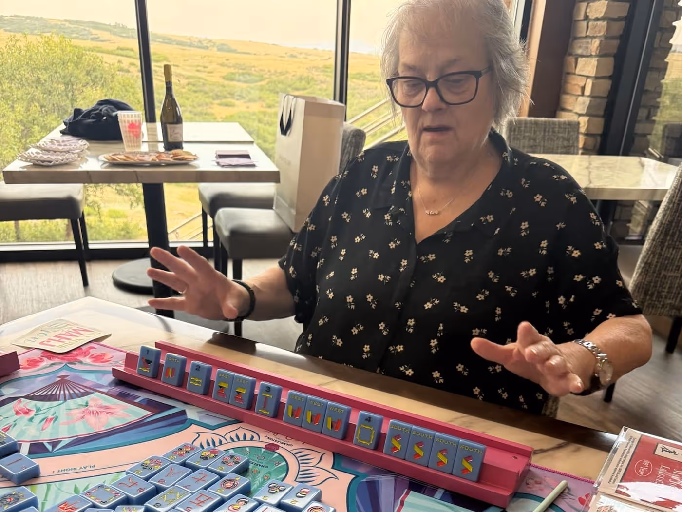 A player at a fully set American mahjong table, hands over her rack of tiles mid-lesson, the floral Mahjong Standard mat and tiles laid out in front of her
