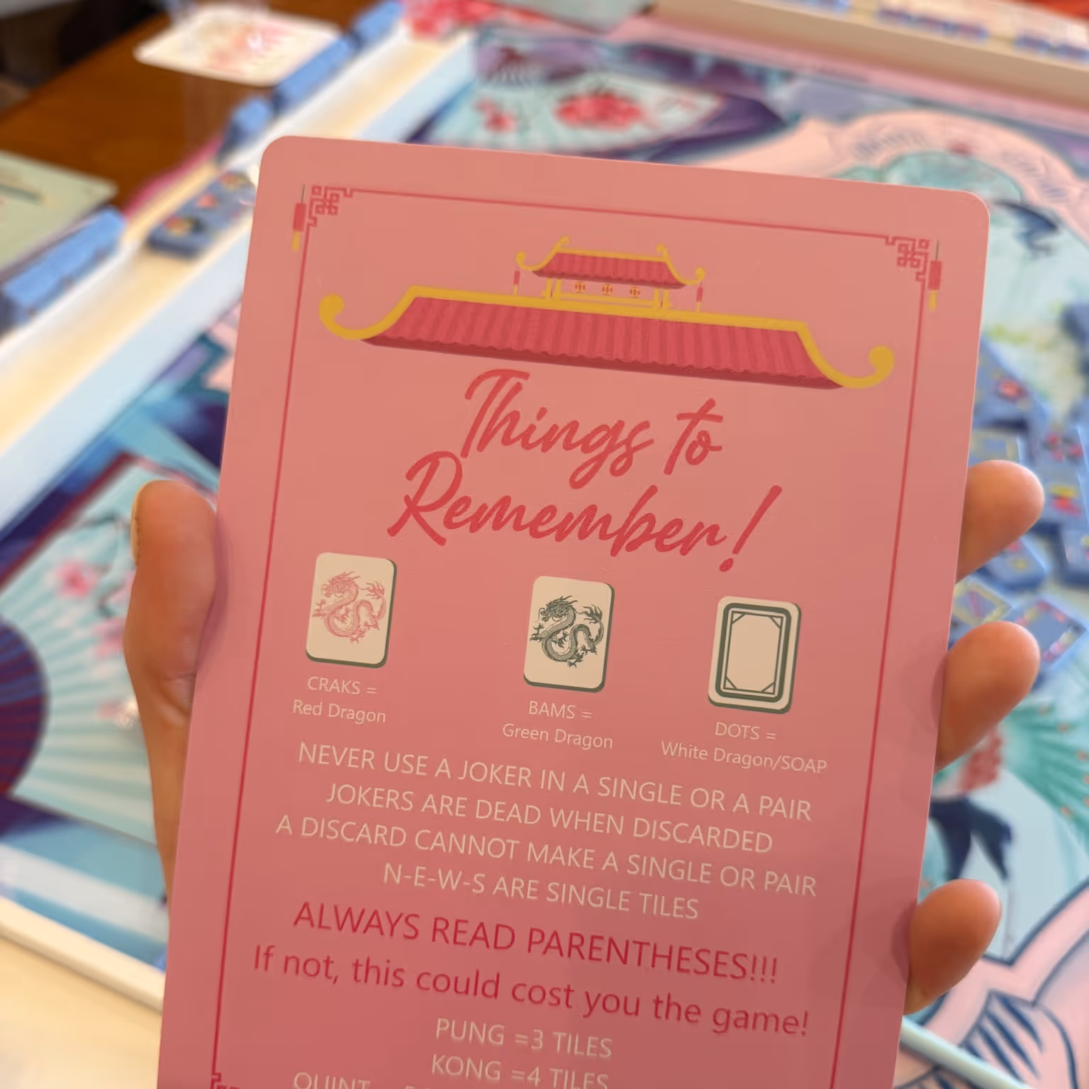 A hand holding Mahjong Standard’s pink “Things to Remember” beginner reference card above a set mahjong table