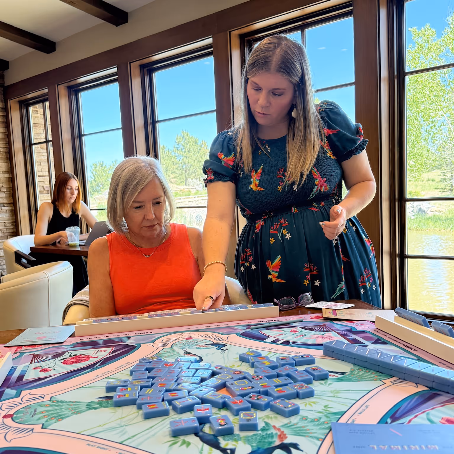 A Mahjong Standard host leaning in to guide a new player through her tiles at a bright, window-lit table in a Highlands Ranch home