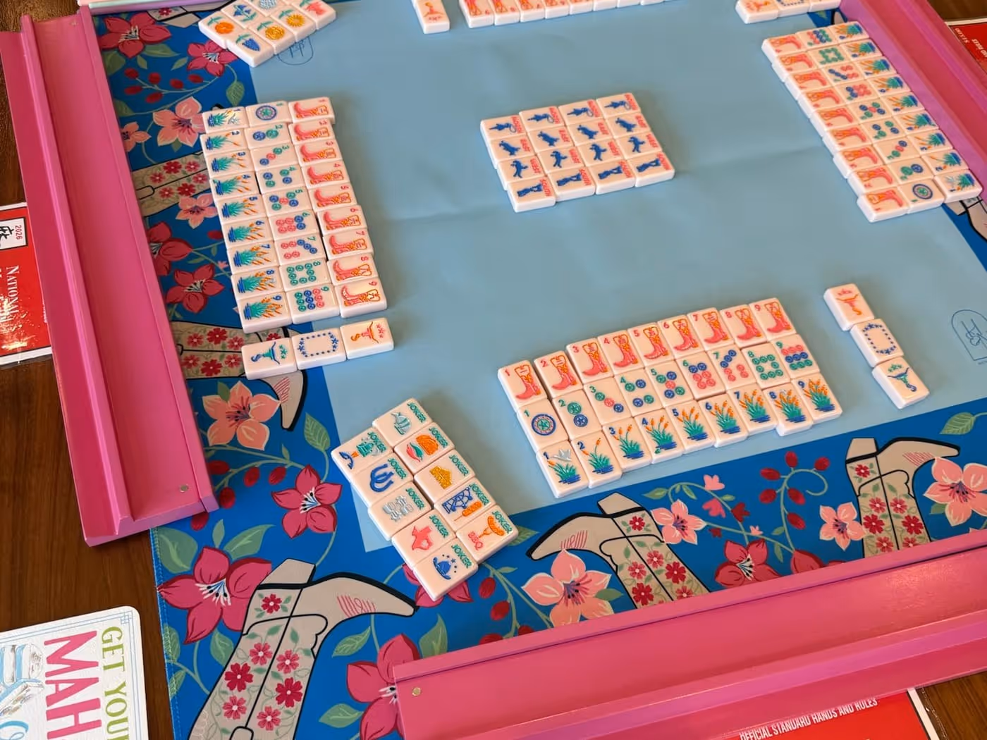 A fully set American mahjong table from above — the pink racks, floral Mahjong Standard mat and tiles laid out, the card and beginner booklet at the edge, ready to play