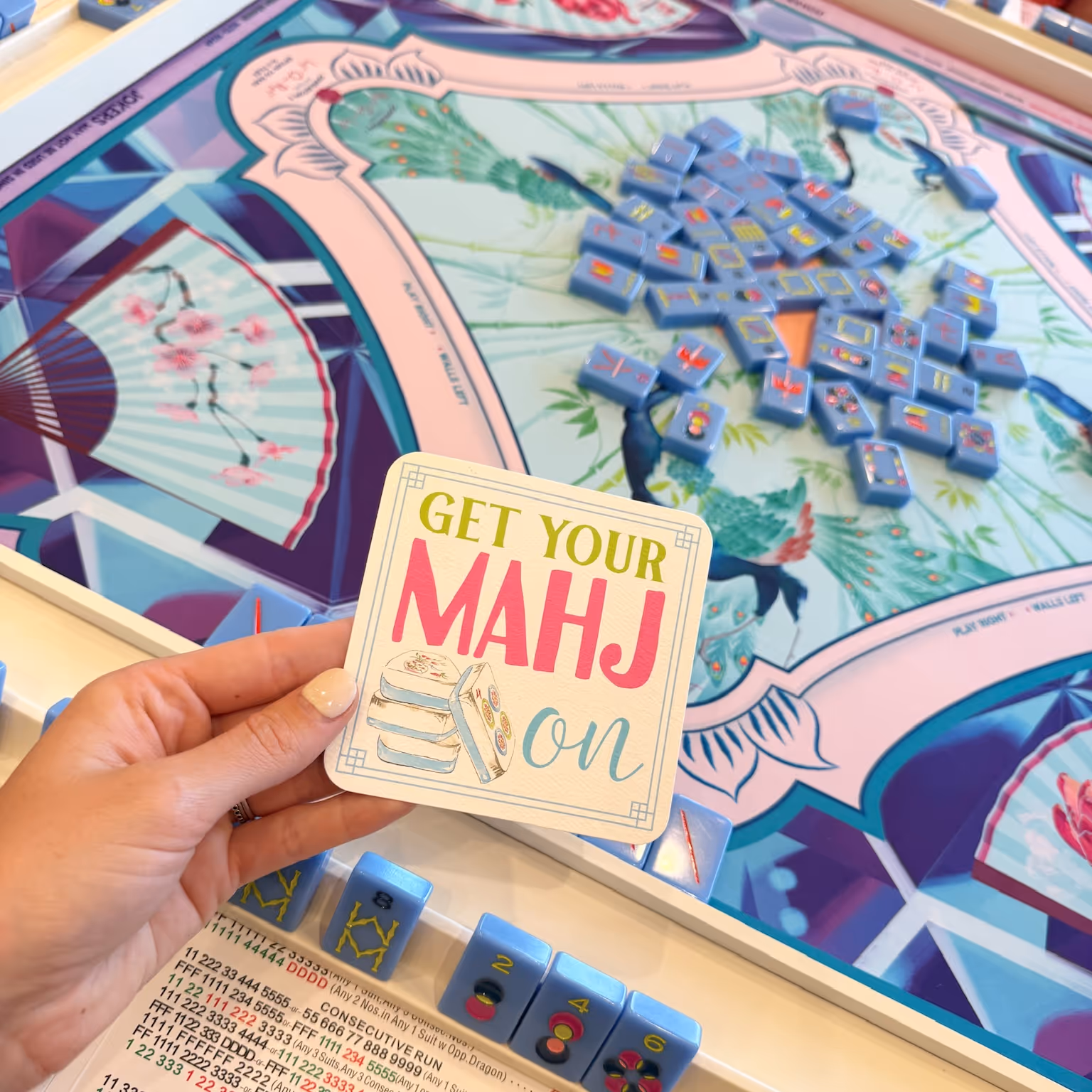 A hand holding a Mahjong Standard “Get your mahj on” card above a colorful playing mat scattered with American mahjong tiles