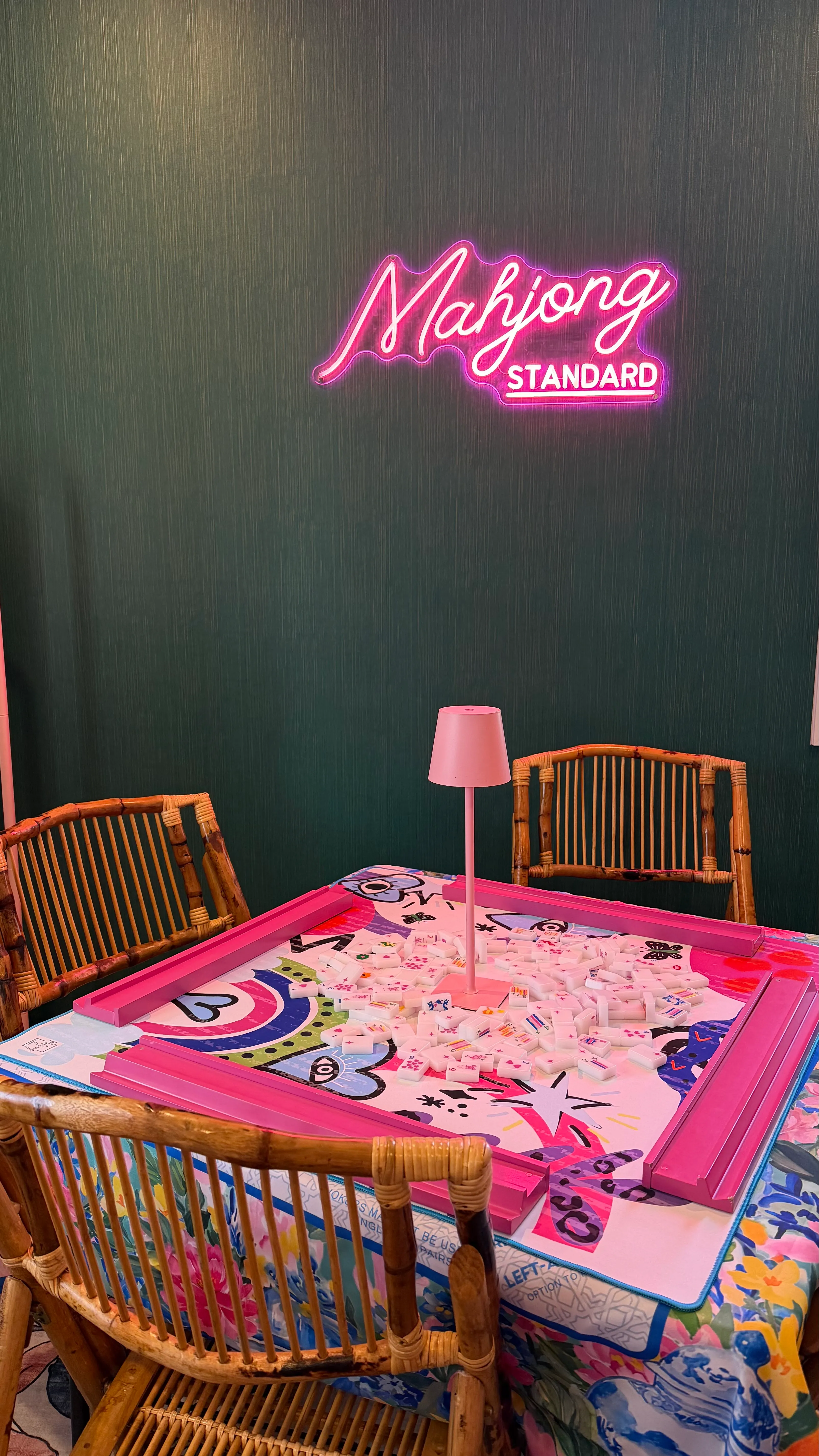 A single mahjong table set for play with a colorful illustrated mat and white tiles, a pink table lamp in the center, beneath a glowing pink Mahjong Standard neon sign on the dark teal wall