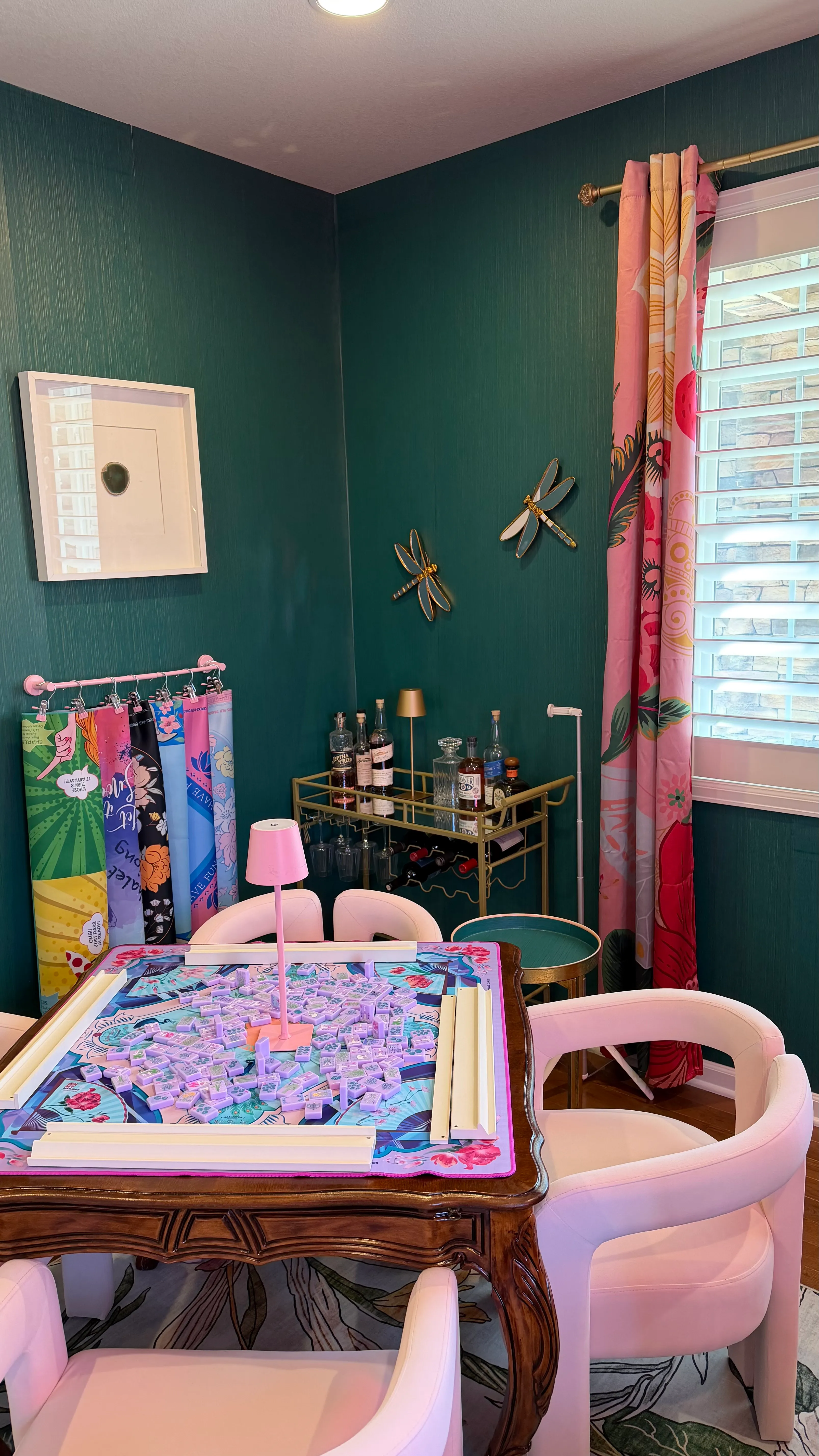 The corner of Courtney's mahjong room in Backcountry, Highlands Ranch: a set mahjong table, pink lamp, vintage bar cart with glassware, and floral curtains beside a window