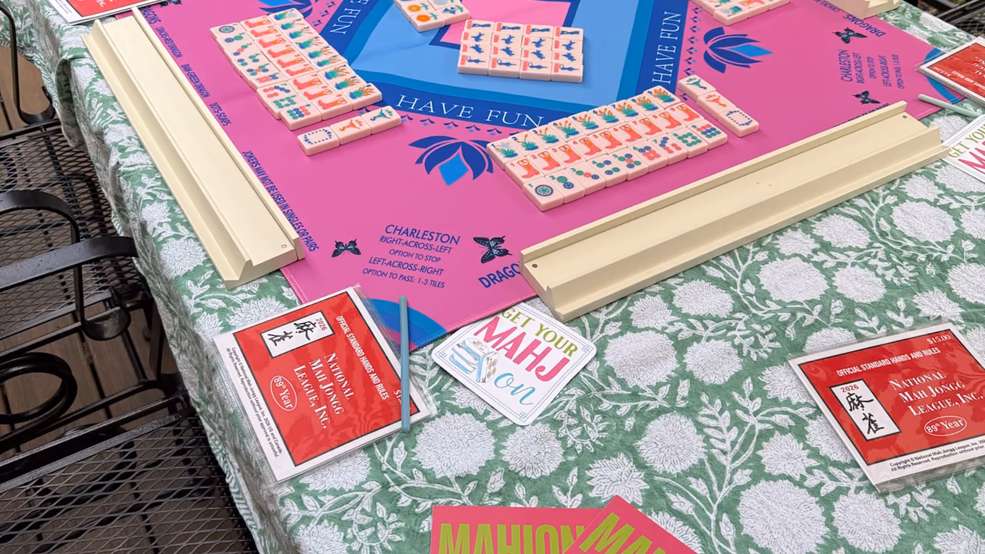 A vibrant pink Mahjong Standard mat with racked colorful tiles and two red National Mah Jongg League card booklets resting on a floral tablecloth, ready to play