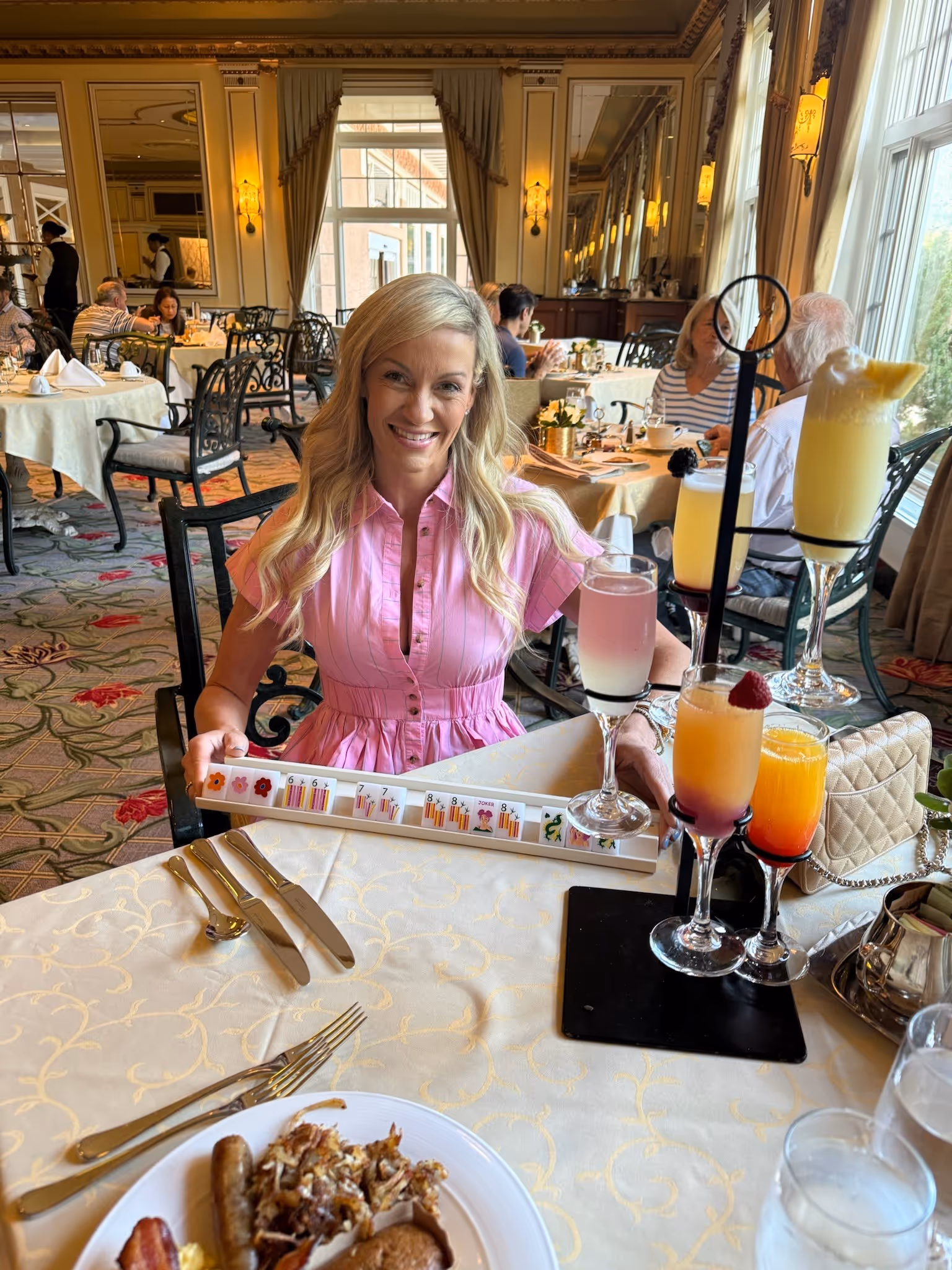 A woman in a pink shirt-dress smiling at a grand hotel brunch, her American mahjong rack in hand and a flight of mimosas beside her