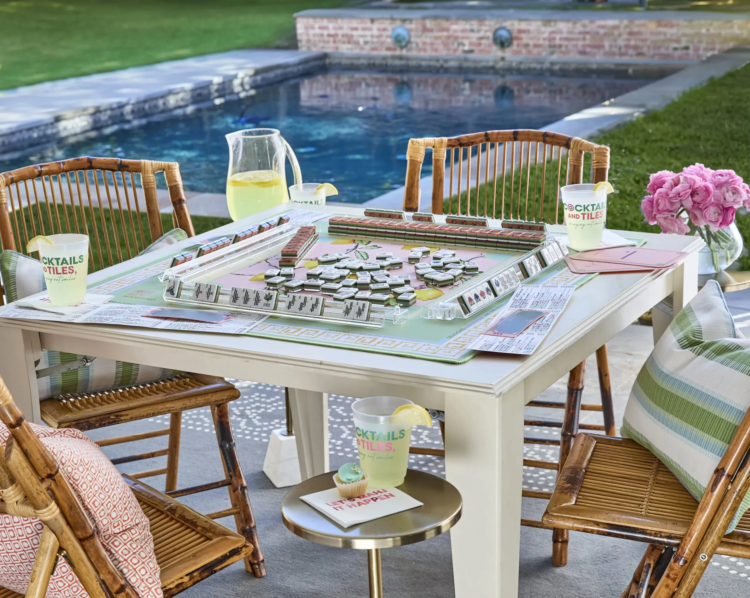 An American mahjong table set for spring play beside a swimming pool, tiles racked and cups on the table in warm afternoon light