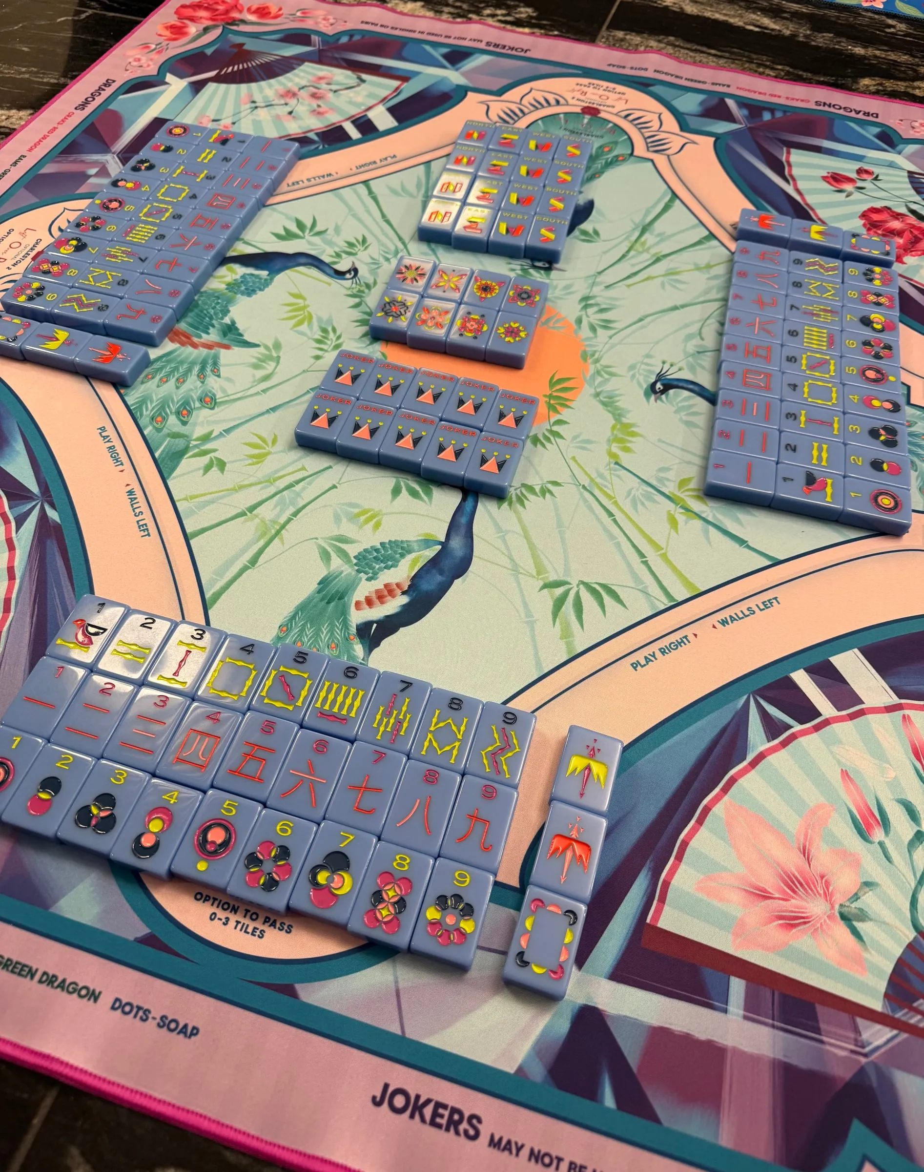 A mahjong game mid-play on a striking peacock-and-bamboo illustrated mat, all four tile walls visible from above, Mahjong Standard tiles in soft blue