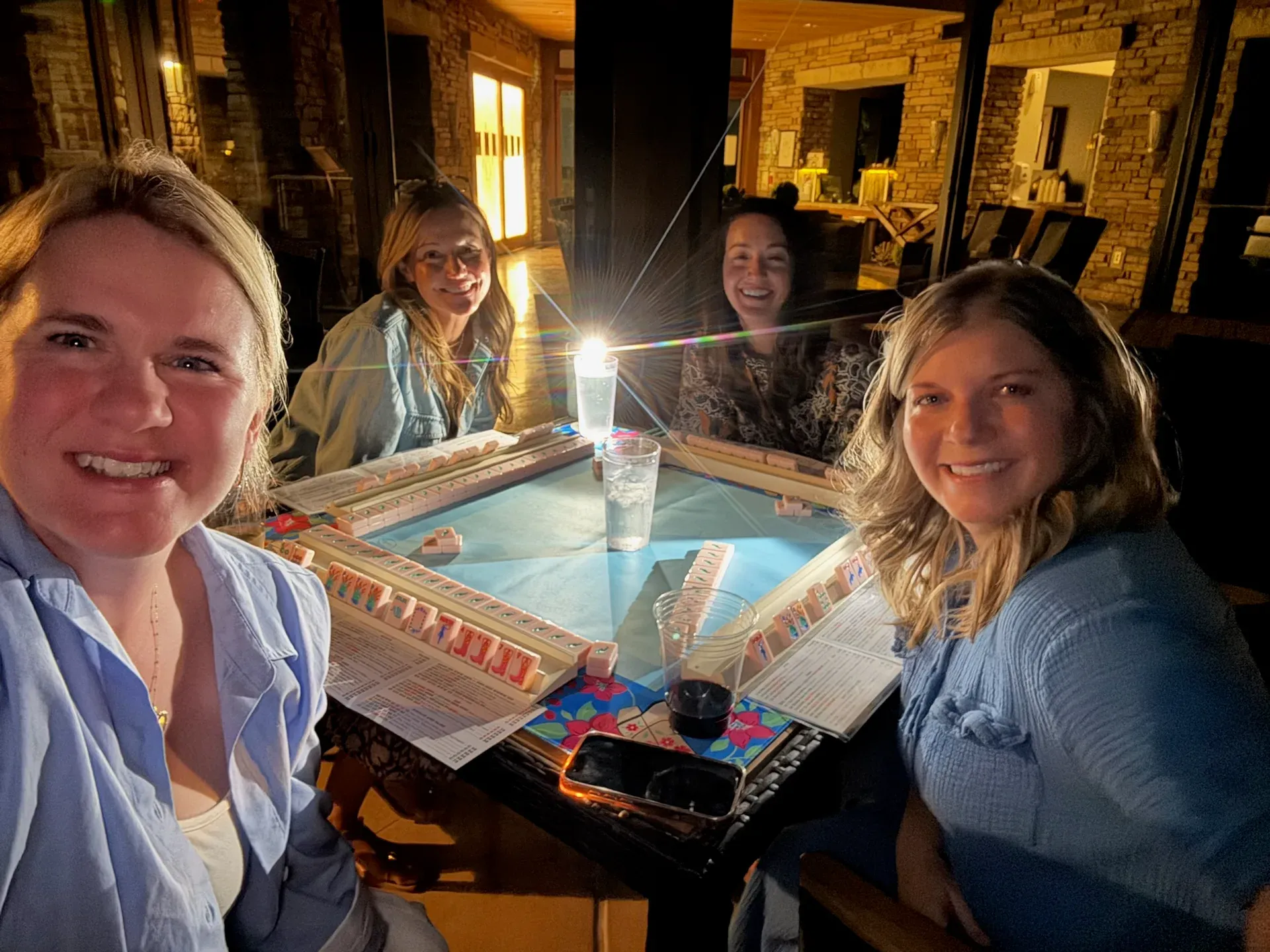 Four friends smiling around a lit mahjong table in the evening, tiles racked and drinks on the table in a warm stone-and-glass room