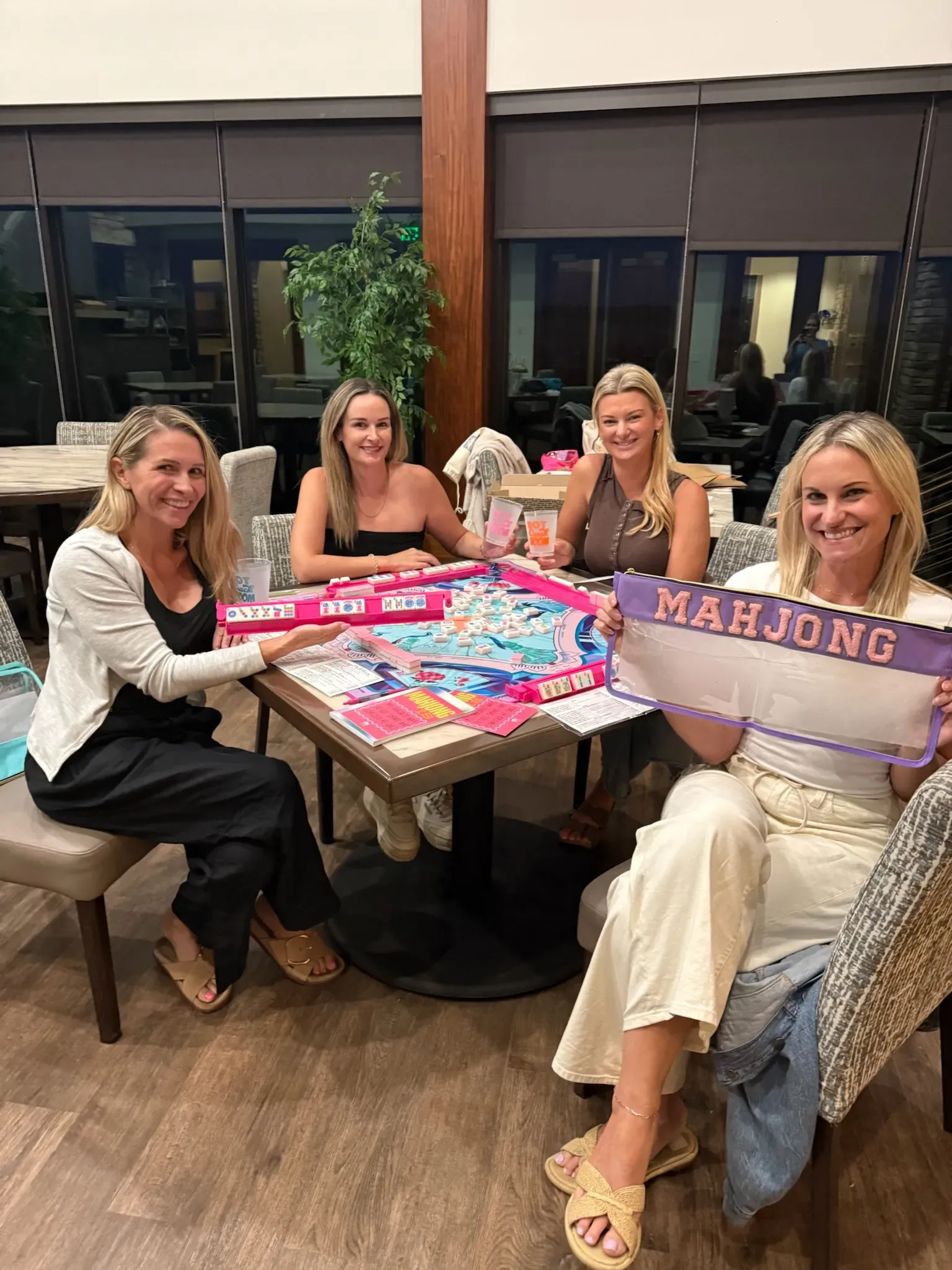 Five friends seated together around a home table with a pink mahjong mat, smiling up at the camera between hands