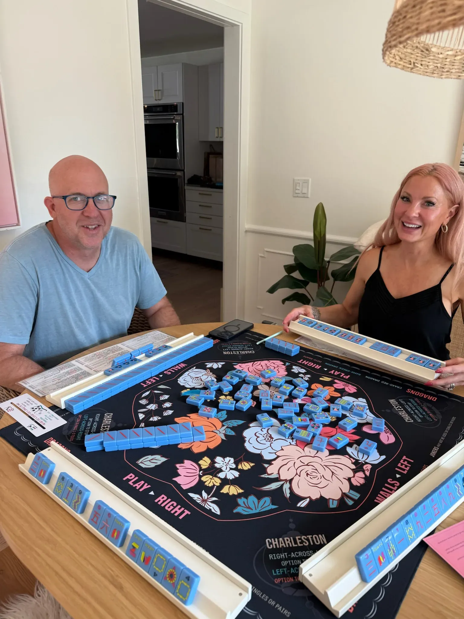 A man and a woman smiling across a mahjong table at home, a floral rose mat between them with blue tiles racked on every side