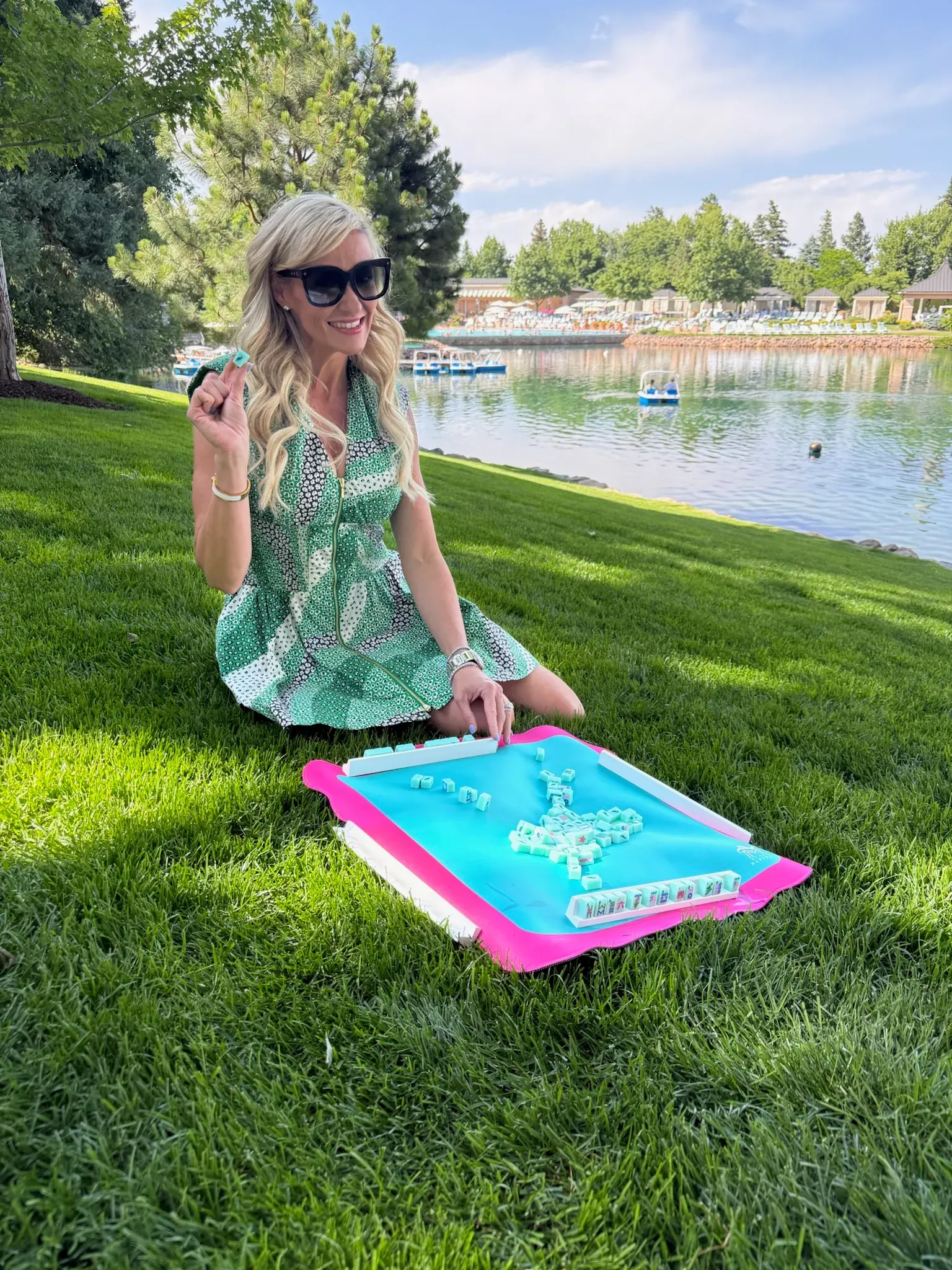 A woman in sunglasses kneeling on the grass by a sunlit pond, smiling as she holds up a bright blue mahjong mat on a summer afternoon