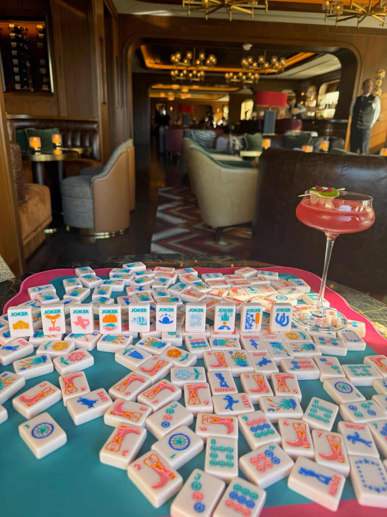 American mahjong tiles spread across the table at Postino with a cocktail beside them, the warm bar lounge behind