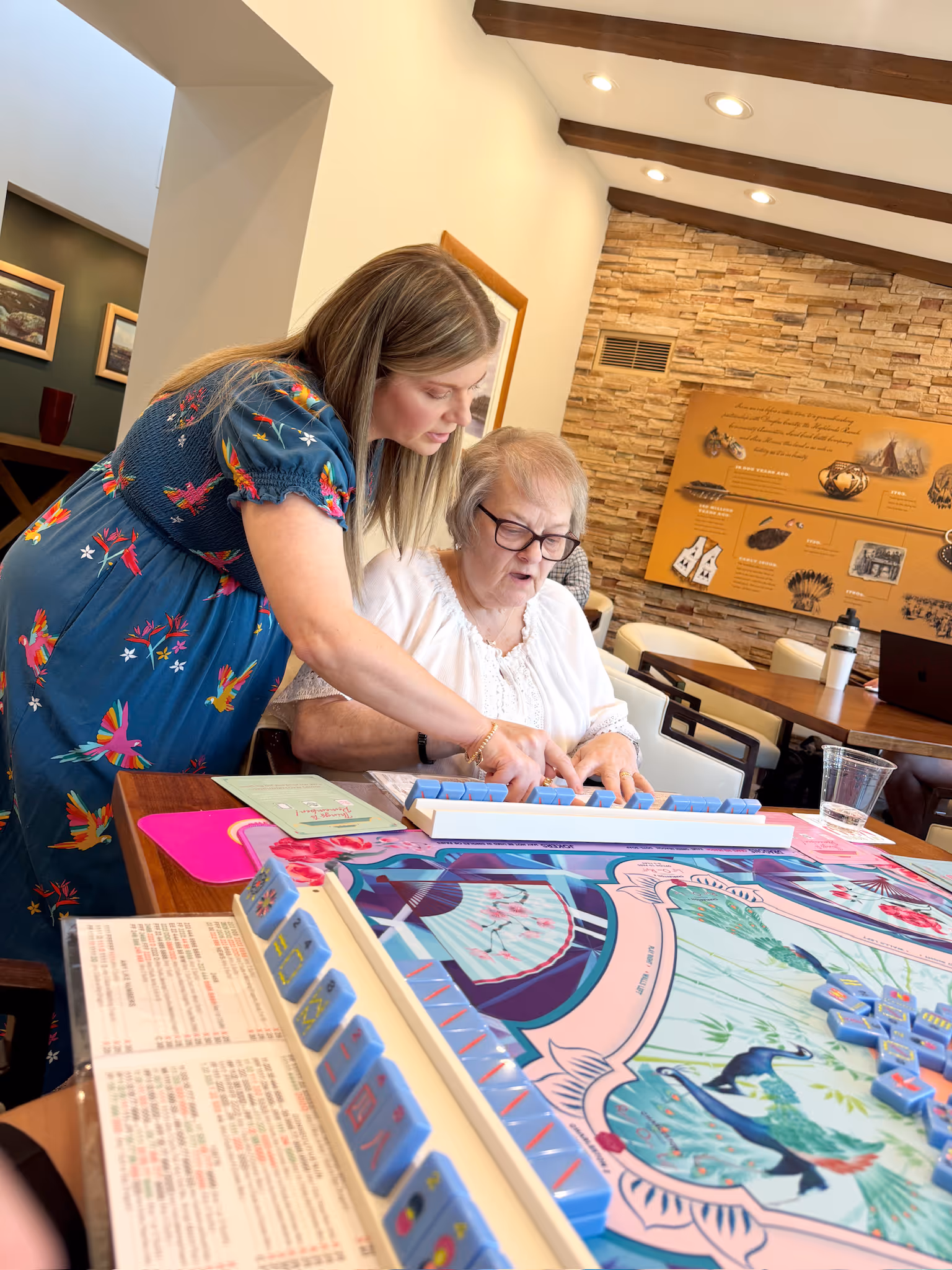 Courtney leaning in to guide a player's hands at the American mahjong table at Postino, tiles laid out on the floral mat between them