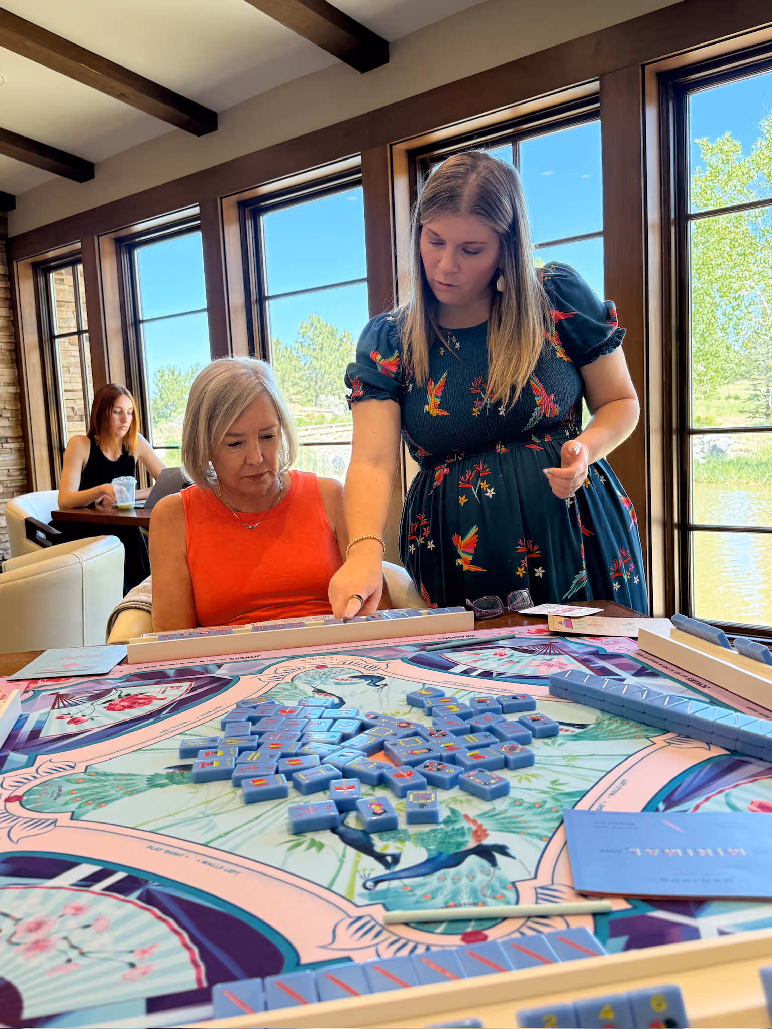 Courtney leaning in to guide a seated player at the American mahjong table at Postino, tiles laid out on the floral mat between them