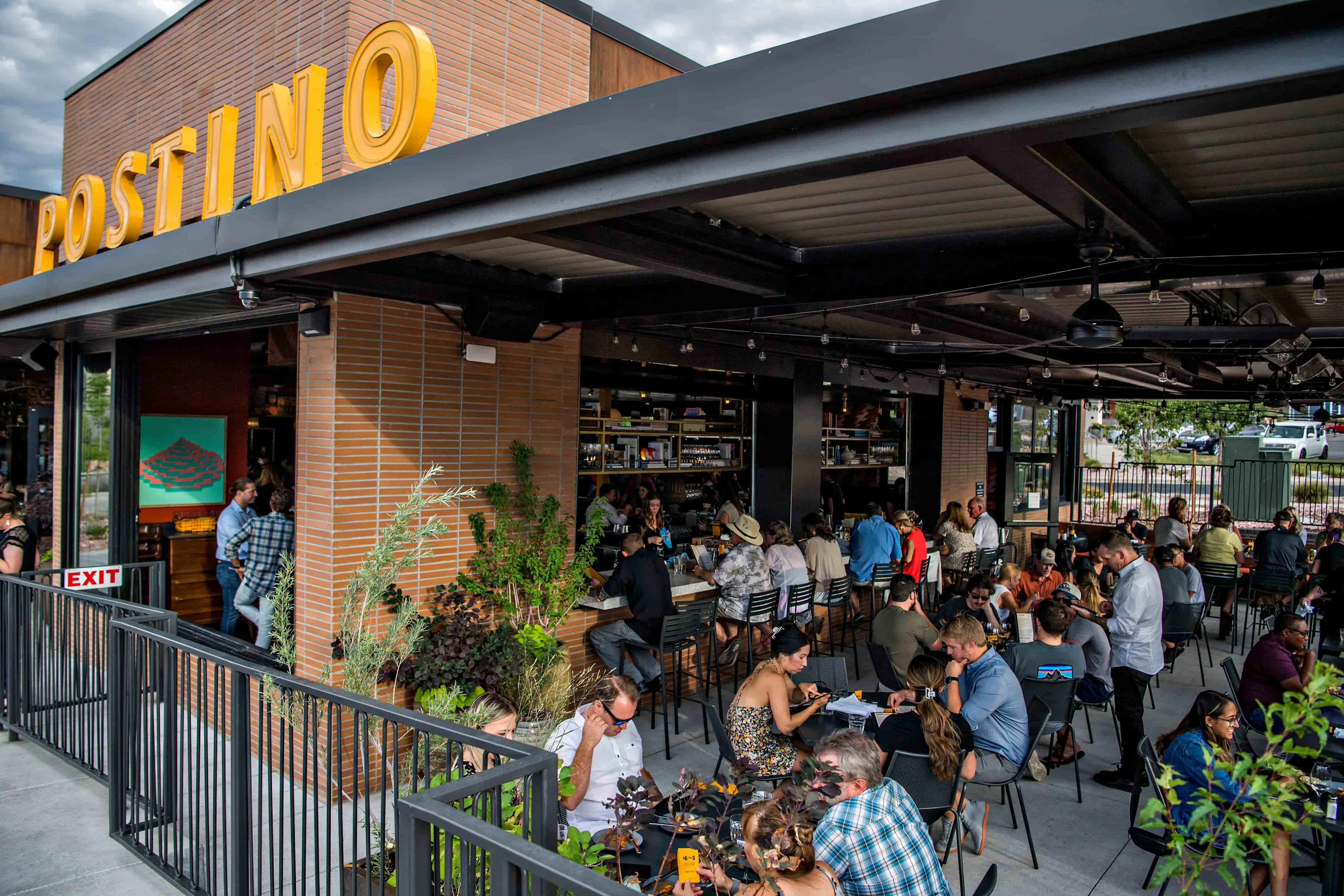 The Postino patio in Highlands Ranch at golden hour, the gold POSTINO wordmark above a busy, buzzing terrace