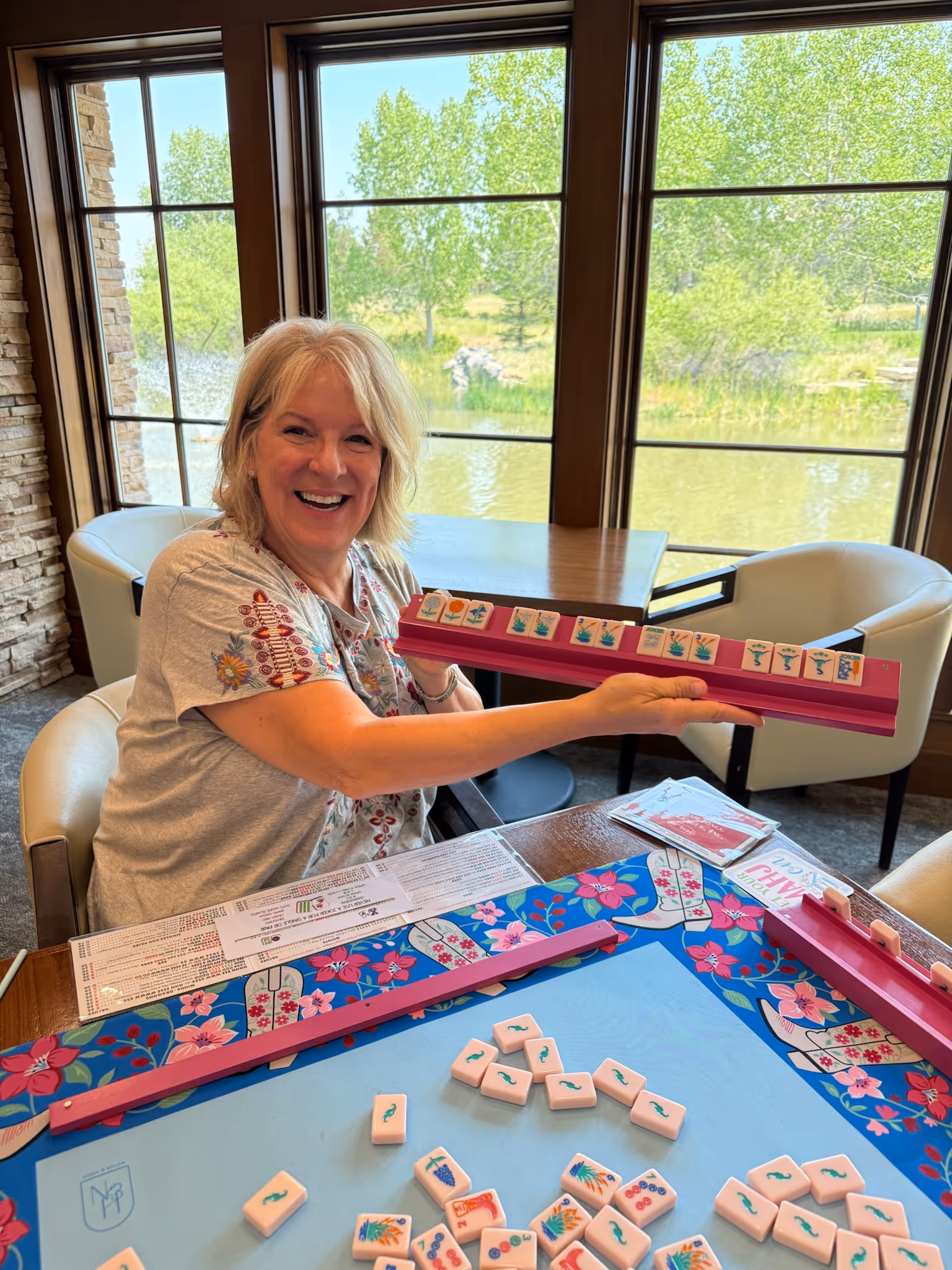 A woman laughing as she holds up her rack of pink American mahjong tiles at Postino, a blue floral mat in front of her and the sunlit windows behind