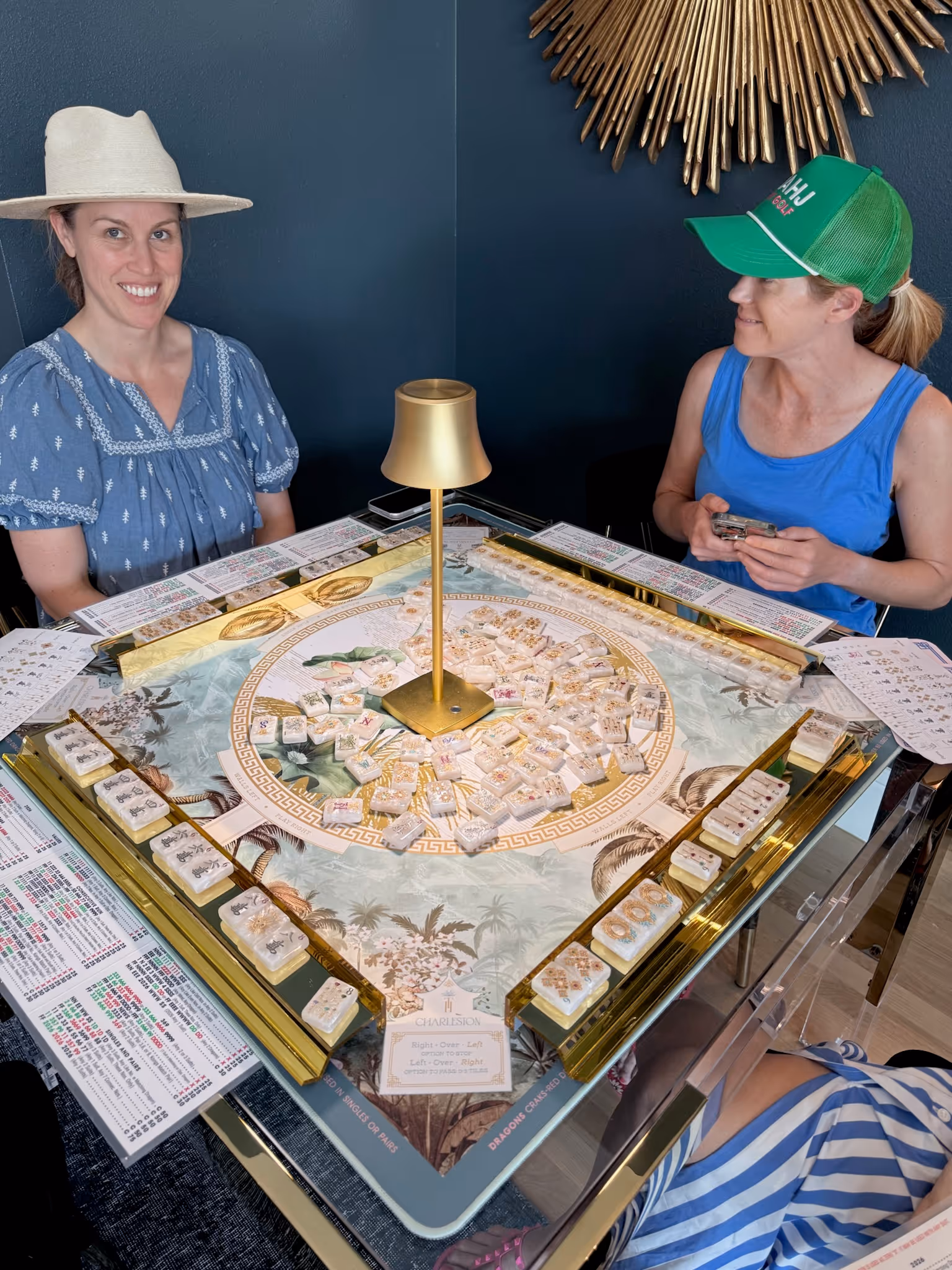 Two women smiling at a glass-and-gold American mahjong table at Postino, a tropical-print mat between them and the tiles racked in walls ready to play