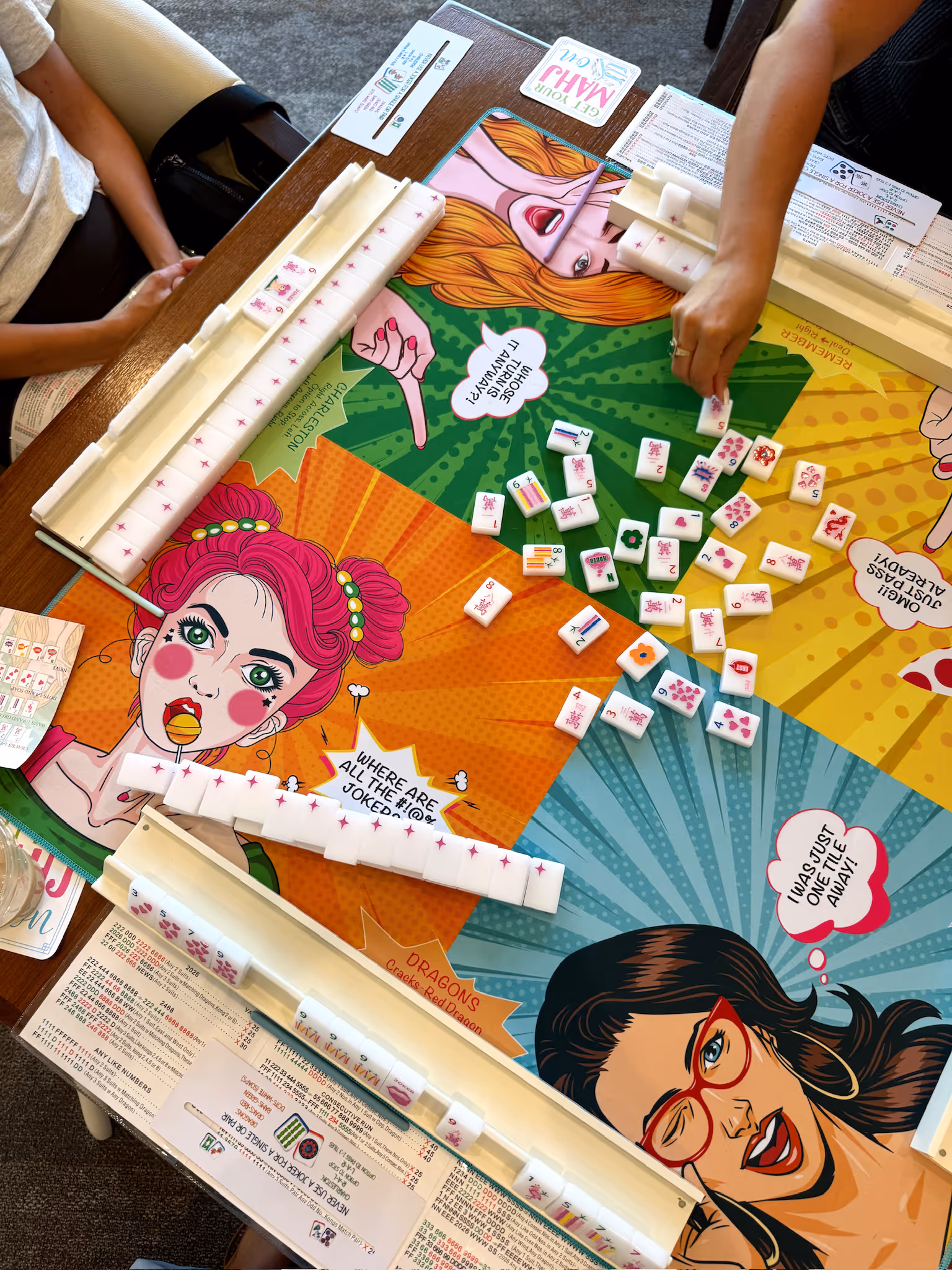 An American mahjong game mid-play seen from above, tiles out across a bright pop-art comic-print mat as players reach in
