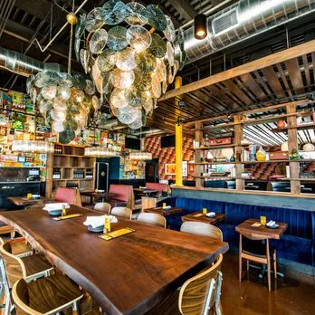 Inside Postino in Highlands Ranch, a long communal wood table flanked by blue banquettes under a shell-disc chandelier, the bar behind
