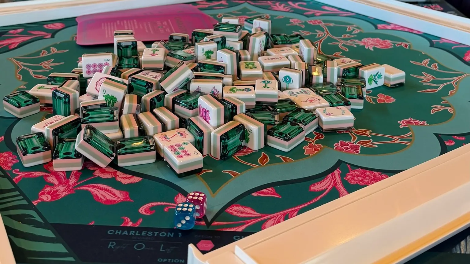 A close-up of emerald green mahjong tiles arranged on a green and pink floral mat, with a pair of blue dice resting alongside