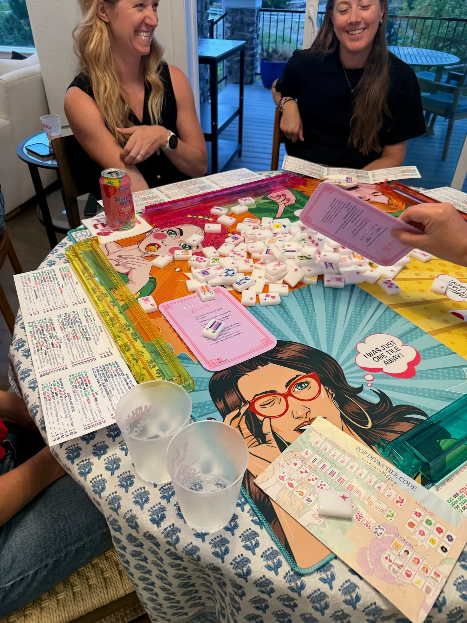 Two friends laughing over an in-home American mahjong game on a colorful illustrated mat, tiles racked and drinks on the table, at a home in Sterling Ranch